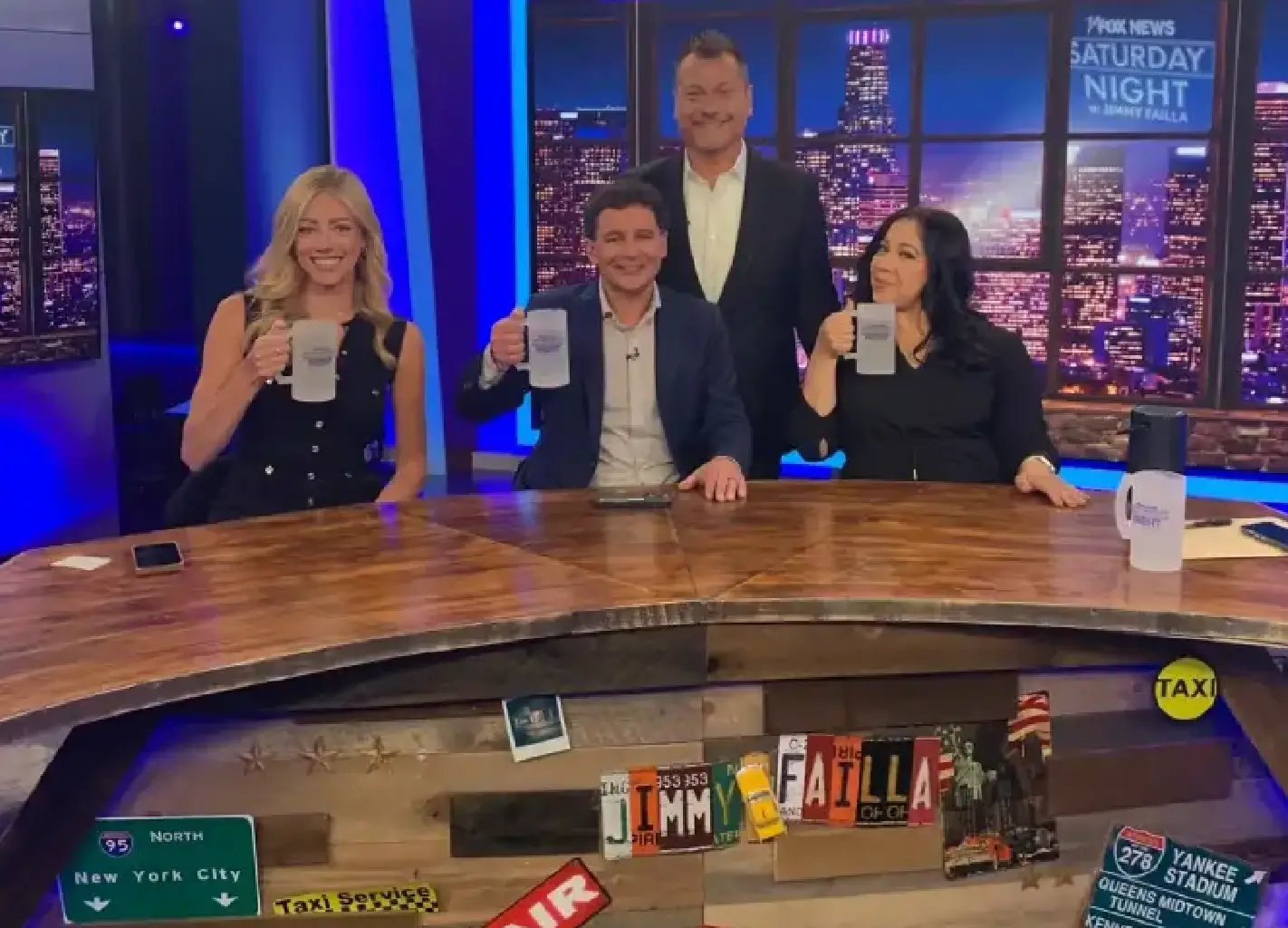 Four people sit at a news desk holding mugs, with a nighttime cityscape in the background and various signs displayed on the front of the desk.